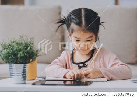 A cheerful Asian mother helps her young daughter do homework and color a book at home, while the father works on a laptop in the background. A cozy, bright, and modern family lifestyle. 136337080