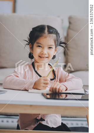 A cheerful Asian mother helps her young daughter do homework and color a book at home, while the father works on a laptop in the background. A cozy, bright, and modern family lifestyle. A cheerful Asian mother helps her young daughter do homework and color a book at home, while the father works on a laptop in the background. A cozy, bright, and modern family lifestyle. 136337082