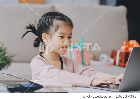 A cheerful Asian mother helps her young daughter do homework and color a book at home, while the father works on a laptop in the background. A cozy, bright, and modern family lifestyle. 136337083