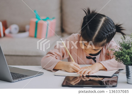 A cheerful Asian mother helps her young daughter do homework and color a book at home, while the father works on a laptop in the background. A cozy, bright, and modern family lifestyle. A cheerful Asian mother helps her young daughter do homework and color a book at home, while the father works on a laptop in the background. A cozy, bright, and modern family lifestyle. 136337085