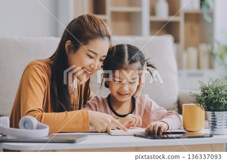 A cheerful Asian mother helps her young daughter do homework and color a book at home, while the father works on a laptop in the background. A cozy, bright, and modern family lifestyle. A cheerful Asian mother helps her young daughter do homework and color a book at home, while the father works on a laptop in the background. A cozy, bright, and modern family lifestyle. 136337093