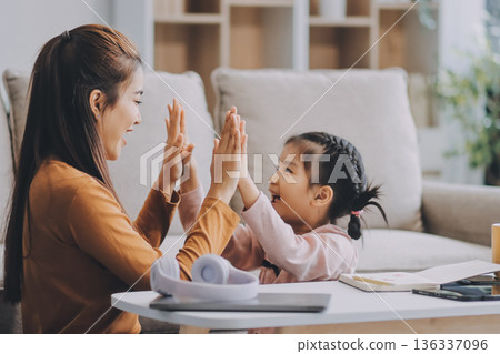 A cheerful Asian mother helps her young daughter do homework and color a book at home, while the father works on a laptop in the background. A cozy, bright, and modern family lifestyle. A cheerful Asian mother helps her young daughter do homework and color a book at home, while the father works on a laptop in the background. A cozy, bright, and modern family lifestyle. 136337096