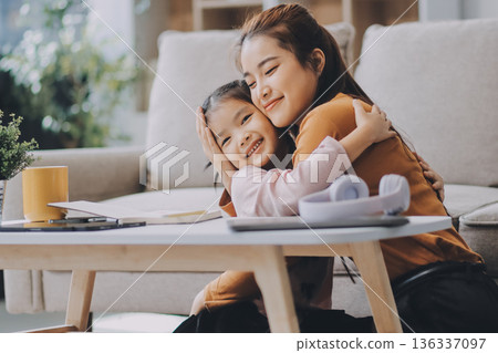 A cheerful Asian mother helps her young daughter do homework and color a book at home, while the father works on a laptop in the background. A cozy, bright, and modern family lifestyle. A cheerful Asian mother helps her young daughter do homework and color a book at home, while the father works on a laptop in the background. A cozy, bright, and modern family lifestyle. 136337097
