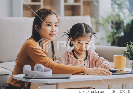 A cheerful Asian mother helps her young daughter do homework and color a book at home, while the father works on a laptop in the background. A cozy, bright, and modern family lifestyle. A cheerful Asian mother helps her young daughter do homework and color a book at home, while the father works on a laptop in the background. A cozy, bright, and modern family lifestyle. 136337098