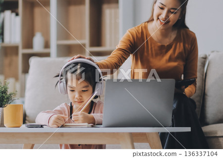 A cheerful Asian mother helps her young daughter do homework and color a book at home, while the father works on a laptop in the background. A cozy, bright, and modern family lifestyle. A cheerful Asian mother helps her young daughter do homework and color a book at home, while the father works on a laptop in the background. A cozy, bright, and modern family lifestyle. 136337104