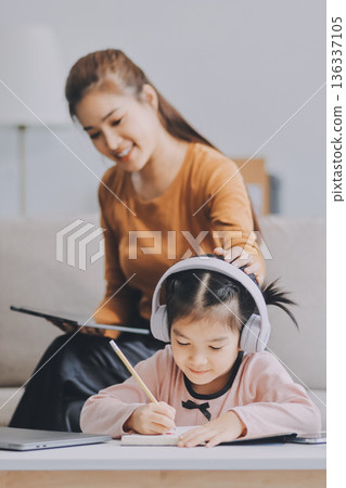 A cheerful Asian mother helps her young daughter do homework and color a book at home, while the father works on a laptop in the background. A cozy, bright, and modern family lifestyle. 136337105