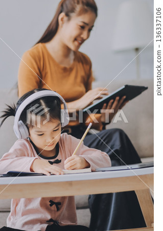 A cheerful Asian mother helps her young daughter do homework and color a book at home, while the father works on a laptop in the background. A cozy, bright, and modern family lifestyle. 136337106