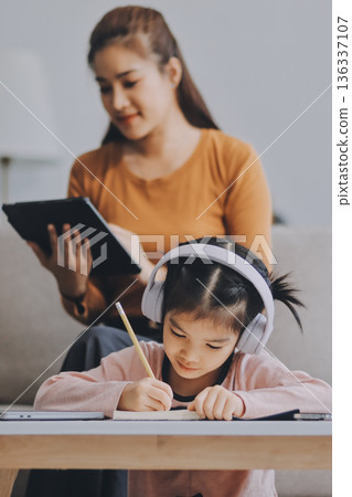 A cheerful Asian mother helps her young daughter do homework and color a book at home, while the father works on a laptop in the background. A cozy, bright, and modern family lifestyle. A cheerful Asian mother helps her young daughter do homework and color a book at home, while the father works on a laptop in the background. A cozy, bright, and modern family lifestyle. 136337107