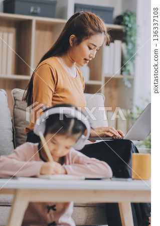 A cheerful Asian mother helps her young daughter do homework and color a book at home, while the father works on a laptop in the background. A cozy, bright, and modern family lifestyle. A cheerful Asian mother helps her young daughter do homework and color a book at home, while the father works on a laptop in the background. A cozy, bright, and modern family lifestyle. 136337108