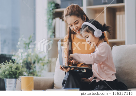 A cheerful Asian mother helps her young daughter do homework and color a book at home, while the father works on a laptop in the background. A cozy, bright, and modern family lifestyle. A cheerful Asian mother helps her young daughter do homework and color a book at home, while the father works on a laptop in the background. A cozy, bright, and modern family lifestyle. 136337115