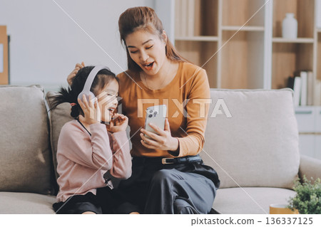 A cheerful Asian mother helps her young daughter do homework and color a book at home, while the father works on a laptop in the background. A cozy, bright, and modern family lifestyle. 136337125