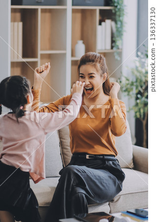 A cheerful Asian mother helps her young daughter do homework and color a book at home, while the father works on a laptop in the background. A cozy, bright, and modern family lifestyle. 136337130