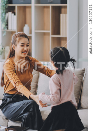 A cheerful Asian mother helps her young daughter do homework and color a book at home, while the father works on a laptop in the background. A cozy, bright, and modern family lifestyle. A cheerful Asian mother helps her young daughter do homework and color a book at home, while the father works on a laptop in the background. A cozy, bright, and modern family lifestyle. 136337131