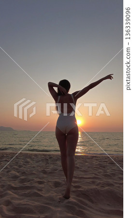 Woman in white swimsuit walking on beach to sunset over sea Woman in white swimsuit walking on beach to sunset over sea 136339096