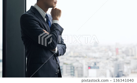 A male business person worries while looking at the city from the office window 136341040