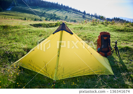 Yellow tourist tent on grassy hillside, accompanied by orange backpack and camera tripod. Stunning view of distant blue mountains under clear sky, perfect for peaceful camping adventure. Yellow tourist tent on grassy hillside, accompanied by orange backpack and camera tripod. Stunning view of distant blue mountains under clear sky, perfect for peaceful camping adventure. 136341674