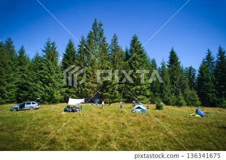 Aerial view of picturesque campsite on grassy hill, surrounded by evergreen trees and overlooking mountain vista. Distant mountains, tourist tents, car, camping gear under golden evening light. Aerial view of picturesque campsite on grassy hill, surrounded by evergreen trees and overlooking mountain vista. Distant mountains, tourist tents, car, camping gear under golden evening light. 136341675