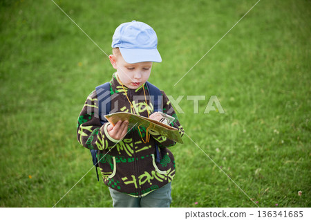 Young boy hiking in grassy field, attentively studying map and compass outdoors. With backpack on shoulders, tourist focused on navigating or planning his route in the mountains. Young boy hiking in grassy field, attentively studying map and compass outdoors. With backpack on shoulders, tourist focused on navigating or planning his route in the mountains. 136341685