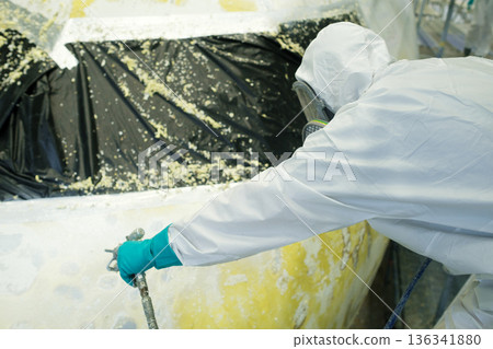 Professional worker applies spray paint to an aircraft in a hangar during repairs, ensuring precision and safety while wearing protective gear 136341880