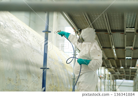 Worker in protective gear spray painting a large surface on scaffolding in an industrial setting during daylight hours Worker in protective gear spray painting a large surface on scaffolding in an industrial setting during daylight hours 136341884