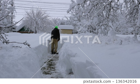 A man in warm clothing, seen from behind, clears a path through high snowdrifts on a residential property leading to a roadway in a rural area. Clearing a path leading from a house to a road A man in warm clothing, seen from behind, clears a path through high snowdrifts on a residential property leading to a roadway in a rural area. Clearing a path leading from a house to a road 136342016