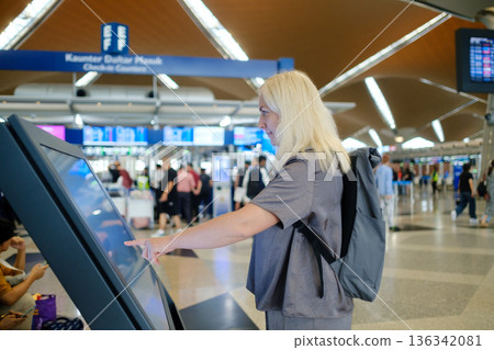 Woman using a touchscreen information kiosk at an airport terminal while travelers wait in the busy background during the daytime Woman using a touchscreen information kiosk at an airport terminal while travelers wait in the busy background during the daytime 136342081
