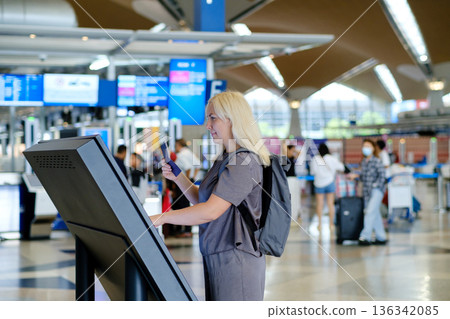 Woman checks in at an airport self-service kiosk while traveling, with busy terminal in the background during afternoon hours Woman checks in at an airport self-service kiosk while traveling, with busy terminal in the background during afternoon hours 136342085