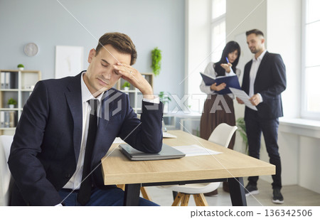 Stressed isolated male employee at desk while colleagues talk in background Stressed isolated male employee at desk while colleagues talk in background 136342506