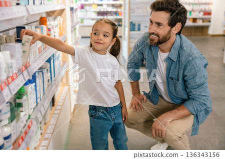 Father squatting next to daughter reaching for product on shelf in pharmacy Father squatting next to daughter reaching for product on shelf in pharmacy 136343156