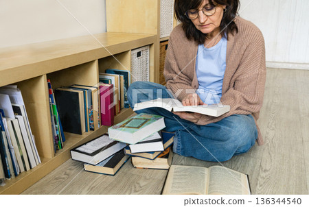 Woman in glasses sitting on floor, learning and studying from books, surrounded by literature at home Woman in glasses sitting on floor, learning and studying from books, surrounded by literature at home 136344540