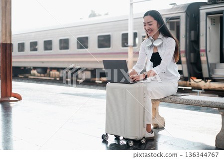 Smiling woman using laptop while sitting on luggage at train station, embracing travel and remote work lifestyle. Smiling woman using laptop while sitting on luggage at train station, embracing travel and remote work lifestyle. 136344767