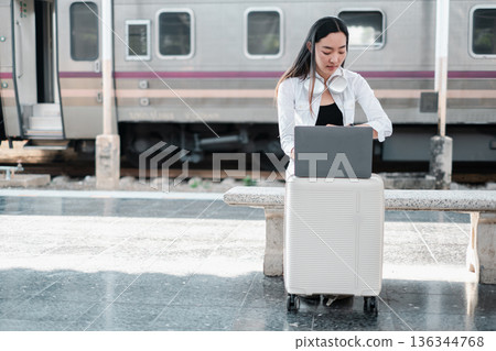 A young woman sits on her suitcase at a train station, working on her laptop, symbolizing travel and remote work. A young woman sits on her suitcase at a train station, working on her laptop, symbolizing travel and remote work. 136344768