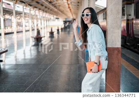 Fashionable woman in sunglasses waves at a train station, showcasing modern travel and style. Fashionable woman in sunglasses waves at a train station, showcasing modern travel and style. 136344771