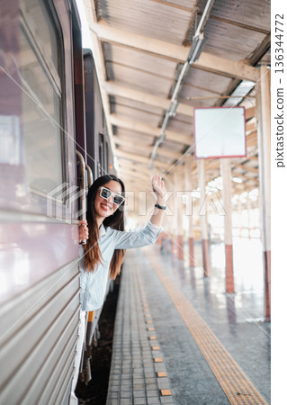 Cheerful woman waving from train window at station, capturing the joy of travel and adventure. Cheerful woman waving from train window at station, capturing the joy of travel and adventure. 136344772