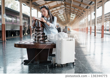 A young woman sits reading at a train station, her luggage beside her, embodying travel and exploration in a modern setting. 136344773
