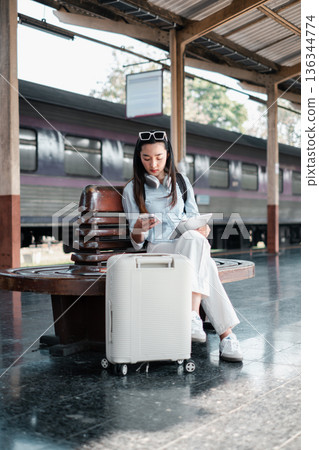 A young woman traveler sits on a bench at a train station with her luggage, using her smartphone and tablet. A young woman traveler sits on a bench at a train station with her luggage, using her smartphone and tablet. 136344774