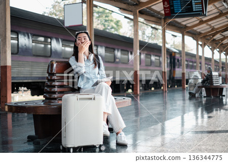 Woman sitting at train station with suitcase, smiling and relaxed, ready for travel adventure. 136344775