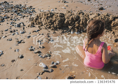 Little girl in a pink swimsuit playing actively with a red toy on the wet sand, building a moat and fort on a sunny pebble beach during a summer vacation Little girl in a pink swimsuit playing actively with a red toy on the wet sand, building a moat and fort on a sunny pebble beach during a summer vacation 136345476