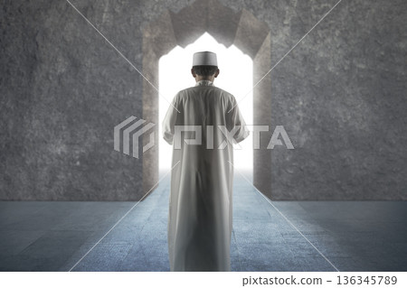 Indonesian southeast asian muslim person wearing traditional white clothes and a white cap, standing and praying facing towards the bright light at a grand stone gateway. Conceptual image of Islam 136345789
