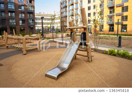 Modern outdoor playground with a metal slide and wooden bridge structure on a safe rubberized surface for child play. Residential area with new architecture. 136345818