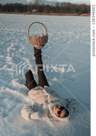 Woman lying in snow with basket on one leg in winter landscape near calm water at sunset Woman lying in snow with basket on one leg in winter landscape near calm water at sunset 136346001