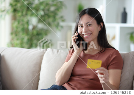 Happy asian woman talking on phone holding card Happy asian woman talking on phone holding card 136346258