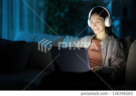 Happy asian woman wearing headphone in the night using laptop 136346263