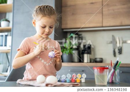 Girl painting pastel-colored eggs while sitting at kitchen table. Calm atmosphere and focused creativity during Easter. 136348198