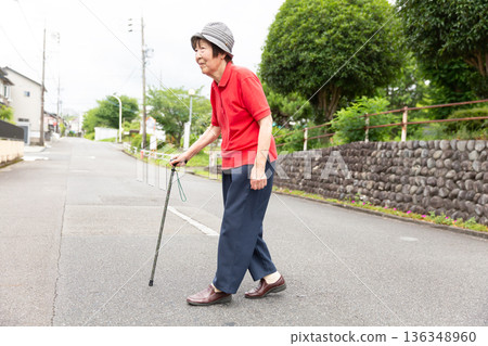 一位老年婦女拄著拐杖在戶外道路行走，全身 136348960