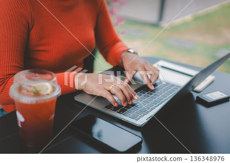 Close-up of Asian woman hands typing on laptop keyboard at outdoor cafe. Freelancer working online with smartphone and iced lemon tea, digital nomad lifestyle and remote work concept. 136348976