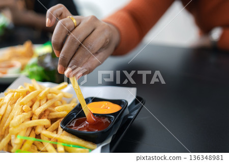 Close up of female hand holding a golden french fry and dipping it into tomato ketchup. Fast food snack on tray with cheese dip. Casual lifestyle eating in a restaurant or cafe background. Close up of female hand holding a golden french fry and dipping it into tomato ketchup. Fast food snack on tray with cheese dip. Casual lifestyle eating in a restaurant or cafe background. 136348981