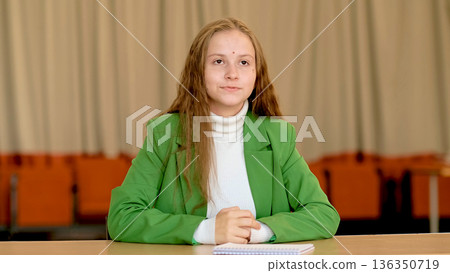 A serious teenage student writes in her notebook at a classroom table, isolated against a soft blurred background and neutral tones. This visual speaks about education systems, personal growth A serious teenage student writes in her notebook at a classroom table, isolated against a soft blurred background and neutral tones. This visual speaks about education systems, personal growth 136350719