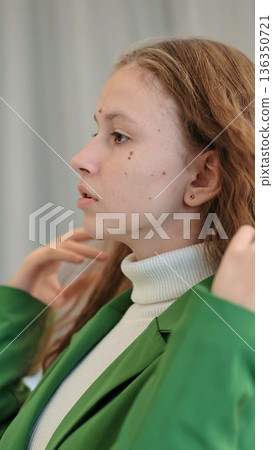 A close-up profile of a young woman adjusting her green blazer, with a contemplative expression highlighting her natural features. The soft background adds to the intimate moment. A close-up profile of a young woman adjusting her green blazer, with a contemplative expression highlighting her natural features. The soft background adds to the intimate moment. 136350721