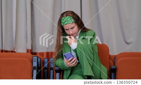 A candid capture of a woman in green sitting in an orange chair, absorbed in her phone. A candid capture of a woman in green sitting in an orange chair, absorbed in her phone. 136350724